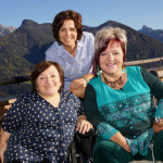 Drei Frauen lächeln am Bergpanorama, inspiriert von einem besonderen Austausch.