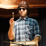 Bärtiger Musiker in Flanellhemd und Sonnenbrille bei Auftritt mit ungewöhnlichem Instrument.