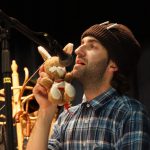 Mann mit Mütze spricht ins Mikrofon und hält ein Plüschhaserl in der Hand.