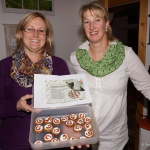 Zwei Frauen halten eine Box mit Keksen und einem Rezept in der Hand, lächelnd und stolz.