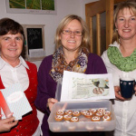 Drei Frauen präsentieren Kekse und Rezeptunterlagen in gemütlicher, ländlicher Umgebung.
