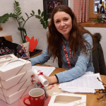 Lächelnde Frau sitzt am Tisch mit Keksen und Unterlagen, bereit für die Suche nach der Kekserlkönigin.