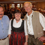 Drei Personen in traditioneller Kleidung lächeln in einem Gasthaus voller Gäste.