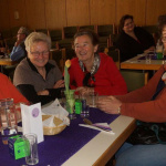 Vier Frauen in bunten Oberteilen lächeln am Tisch, umgeben von Getränken und Snacks.