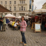 Wochenmarkt mit Ständen und Besuchern, im Vordergrund eine Frau mit Getränk.