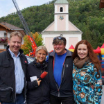 Vierte Personen posieren vor der Kirche in Lend, umgeben von Herbstfarben und Spielgeräten.
