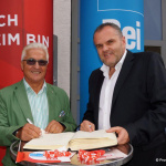 Zwei Männer stehen an einem Tisch und schreiben in ein Buch, mit rednerischen Plakaten im Hintergrund.