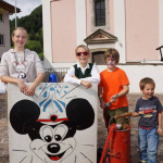 Kinder posieren vor einem Mickey-Mouse-Schild in Lend, idyllische Kulisse im Hintergrund.