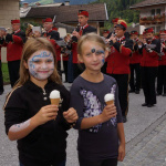 Zwei Kinder mit Eis und Gesichtsbemalung vor einer Feier mit Musikkapelle in Lend.