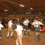 Tanzende Männer in Tracht vor begeistertem Publikum bei einer regionalen Veranstaltung.